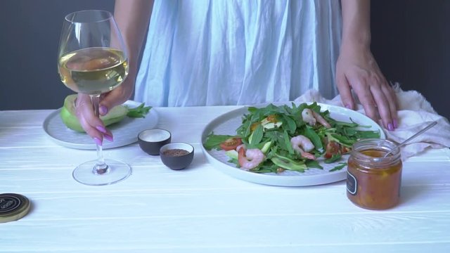 A woman on a table with a salad, a can of sauce and avocado in a plate fills a glass of white wine