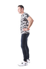 Naklejka premium Side view of young masculine thoughtful man in army t-shirt with hands on hips looking away. Full body isolated on white background. 