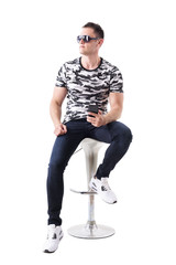 Cool man wearing sunglasses and camouflage t-shirt holding mobile phone looking away sitting on bar stool. Full body isolated on white background. 