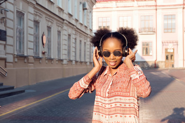 Black afroamerican girl has a walk in evening town and listening to music in headphones.  Girl in sunglasses in summer smiles and looking on sunset.