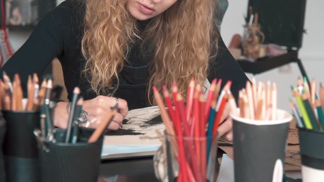 Professional young artist drawing a picture in the sketchbook, working at the desk with lots of various pencils, has lovely tattoo of flower on right hand