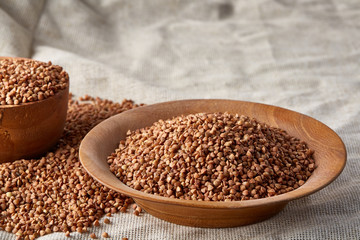 Bowl and plate full of buckwheat grains on homespun tablecloth, close-up, selective focus, shallow depth of field.