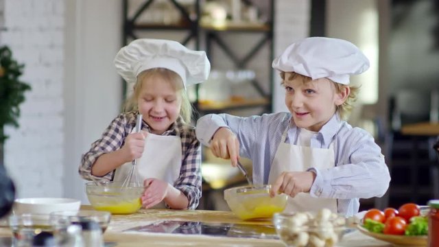 Two adorable little children in aprons and chef hats beating eggs with whisks and talking when cooking in kitchen together