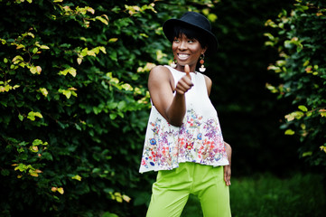 Amazing african american model woman in green pants and black hat posed with different emotions at park, shows a thumb up.