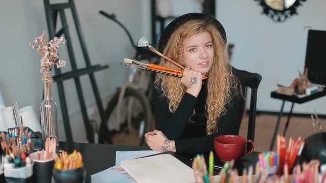 Smiling artist holding paintbrushes, sitting at black desk full of pencils, having a break before creating another masterpiece, portrait