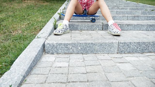 Pan up girl with smartphone in hands playing a game on it while sitting on her skateboard on the stairs in the park. Slow motion footage