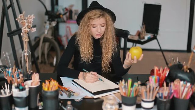 Good-looking artist drawing an apple, trying to delineate yellow fruit into the sketchbook, having lovely tattoos on the hands