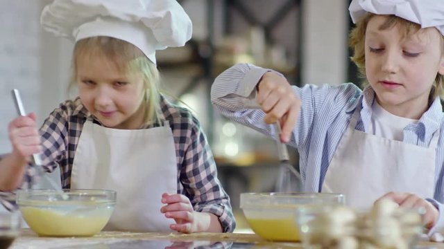 Medium shot of two cute children in chef hats and aprons learning to cook and whisking eggs in glass bowls