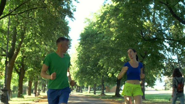 Cheerful female athlete runner overtaking male jogger on park trail and sticking out her tongue during running in nature. Playful brunette sporty fit woman outrunning male jogger during run in park.