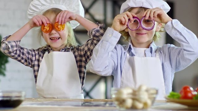 Medium shot of two adorable children in aprons and chef hats holding onion and tomato slices over eyes and looking at camera