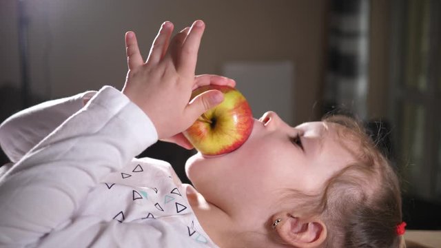 Little funny child girl at home eating biting organic fruit ripe apple lying on the table