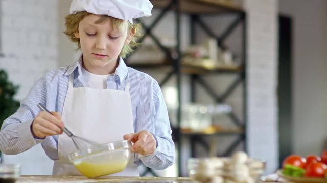 Medium shot of cute little boy in apron and chef hat looking at camera and beating eggs with whisk