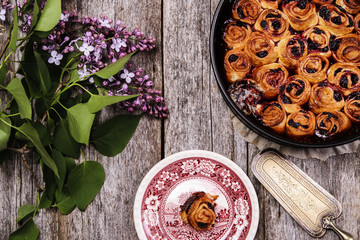 Homemade mini cherry rolls in a baking form and one roll on a plate with bouquet of lilac on vintage wooden table. Bun cake with cherry filling