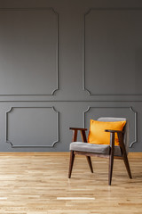 Orange pillow on wooden armchair in simple grey loft interior with wall with molding. Real photo