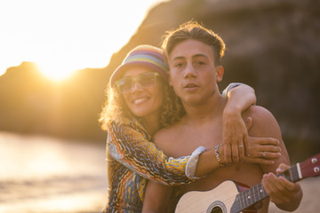 family leisure activity at the beach during a golden colored sunset in vacation and freedom lifestyle. mother and son together hugging and playing music with acoustic guitar. happiness concept