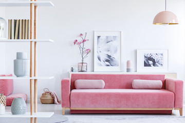 Real photo of a pink couch with pillows in front of a shelf with posters, candle and flowers in bright living room interior with a hanging lamp and shelf with books in the front