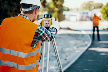 Engineer in orange reflective vest using professional equipment during carriageway work - obrazy, fototapety, plakaty