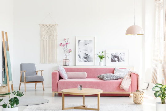Real photo of a pink couch with pillows standing behind a table, next to an armchair, in front of a shelf with posters and flowers and under a lamp in bright and cozy living room interior © Photographee.eu