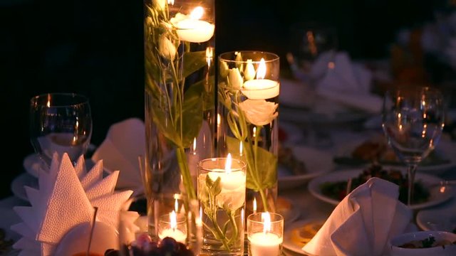 Wedding banquet hall interior details with decorated table setting at restaurant. Candles and white petals decoration with roses flowers in glass vases filled with water.