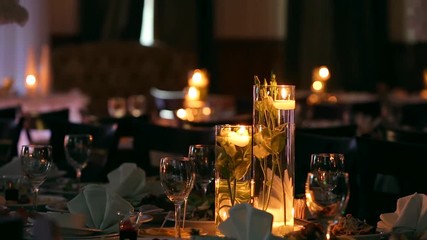 Wedding banquet hall interior details with decorated table setting at restaurant. Candles and white petals decoration with roses flowers in glass vases filled with water.