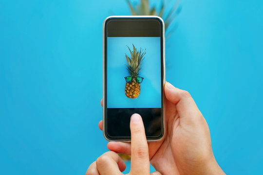 instagram fruit photo. hands holding phone and taking photo of pineapple in sunglasses on blue paper, trendy flat lay. stylish food photography. creative phone image