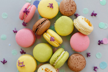 colorful macarons on trendy pastel gray paper with lilac flowers and confetti, flat lay. tasty pink, yellow, green and brown macaroons. candy bar for party. food photography