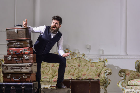 Man with beard and mustache packed luggage, white interior background. Move out, transportation concept. Macho elegant on desperate face, exhausted at end of packing, near pile of vintage suitcases.
