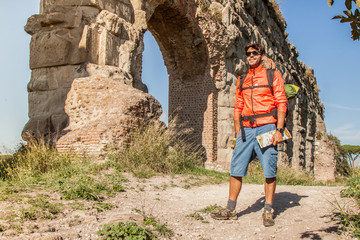 Backpacker posing under majestic ancient roman aqueduct at sunset. Young attractive athletic man with orange sportswear and backpack in parco degli acquedotti in Rome