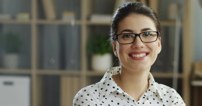 Close up of the Caucasian young beautiful woman in the white blouse and glasses working at the computer in the office and smiling. Portrait. Indoor