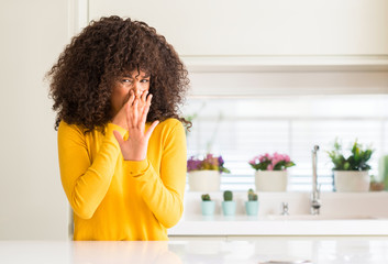 African american woman wearing yellow sweater at kitchen smelling something stinky and disgusting,...