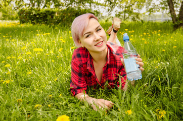 Young beautiful girl lies on a green grass and dandelions, holds bottle of water.