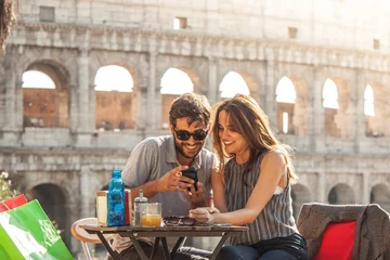 Wall murals Rome Happy young couple tourists using smartphone sitting at bar restaurant in front of colosseum in rome at sunset with coffee shopping bags smiling having fun texting browsing and sharing pictures