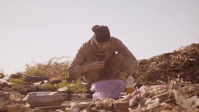 dirty homeless hungry man in a dump eating orange for food in the package with walking goes looking for food slow motion video. homeless dirty man roofless person looking for food in a dump. refugee