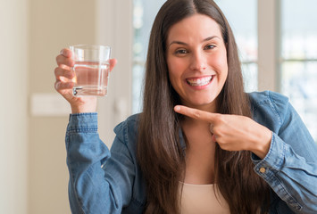 Young woman drinking glass of water at home very happy pointing with hand and finger