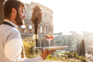 Elegant waiter holding a serving try with a glass of colorful icecream in front of colosseum in rome at sunset