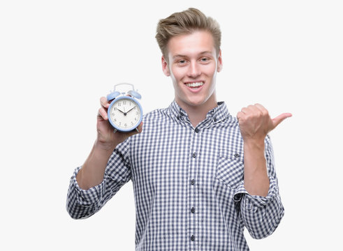 Young handsome blond man holding alarm clock pointing with hand and finger up with happy face smiling