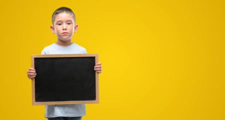 Dark haired little child holding a blackboard with a confident expression on smart face thinking...