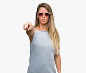 Beautiful young woman wearing sunglasses and denim dress pointing with finger to the camera and to...