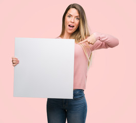 Beautiful young woman holding advertising banner very happy pointing with hand and finger