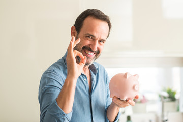 Middle age man save money on piggy bank doing ok sign with fingers, excellent symbol