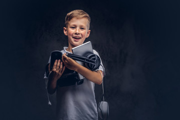 Joyful Cute schoolboy dressed in a white t-shirt, holds full digital set for entertainment at a studio. Isolated on dark textured background.