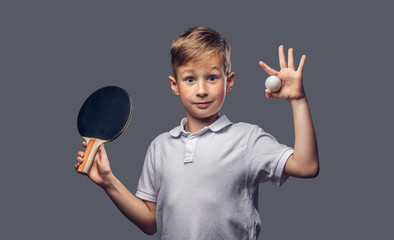 Redhead schoolboy dressed in a white t-shirt holds a ping-pong racquet and ball in a studio....