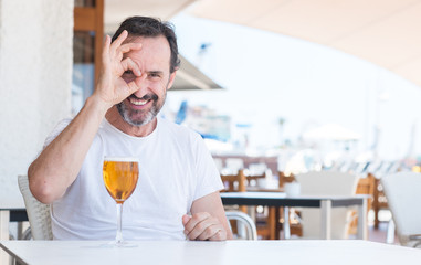 Handsome senior man drinking beer at restaurant with happy face smiling doing ok sign with hand on eye looking through fingers