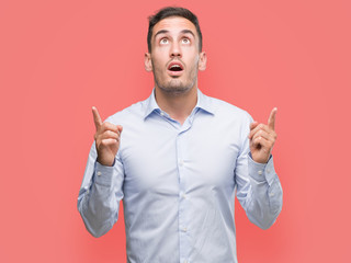 Handsome young businessman amazed and surprised looking up and pointing with fingers and raised...