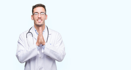 Handsome young doctor man praying with hands together asking for forgiveness smiling confident.