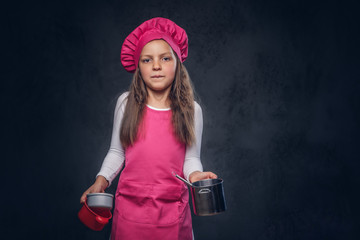 Cute beautiful schoolgirl dressed in a pink cook uniform holds cookware at a studio. Isolated on a dark textured background.
