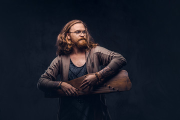 Fototapeta na wymiar Redhead hipster male with long luxuriant hair and full beard dressed in casual clothes playing on a Russian traditional musical instrument - gusli. Isolated on a dark background.
