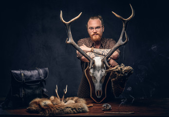 Redhead taxidermist hipster male in sunglasses dressed in a brown shirt, standing near a table with...