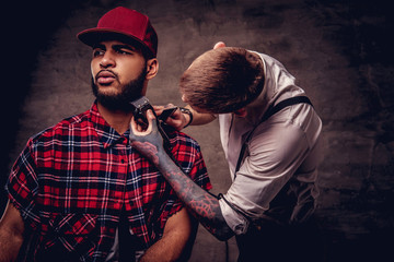 Handsome bearded African American guy in a fleece shirt and cap getting a haircut by an old-fashioned professional hairdresser does haircut.