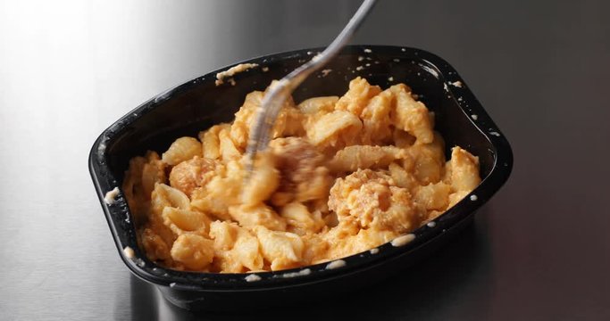 Close video of a spicy chicken nuggets with macaroni and cheese TV dinner in a black tray on a stainless steel counter top being eaten.
